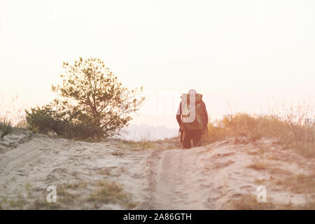 Abbildung der männlichen Backpacker Reisenden gehen auf sandigen Weg während des Sonnenuntergangs. Ein Mann Spaziergänge entlang der sandigen Weg. Stockfoto