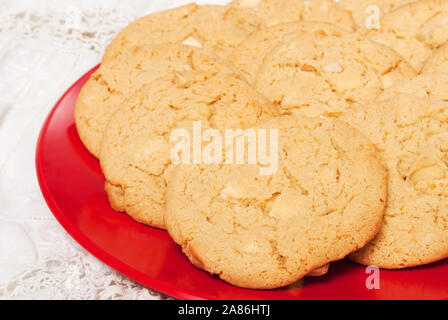 Weiße Schokolade Macadamianuss Cookies serviert auf einem rotem Kunststoff Platte mit einem antiken Bettwäsche Tischdecke als Hintergrund. Fokus auf den Vordergrund schafft Sh Stockfoto