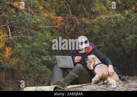 Erfahrene Reisende mit Laptop mit Freund pet während des Gehens Reise in den Wald. Mann umarmt einen Hund Stockfoto