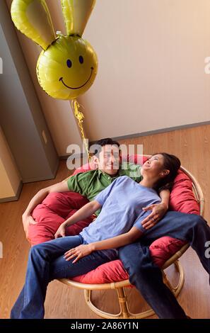 Asiatische amerikanische Paar heraus zusammen hängen in einem großen Sessel mit einem dummen Ballon über schweben Stockfoto