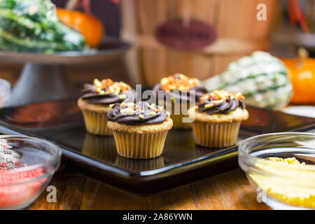 Herbst themed Mini Muffins. Yellow Cake mit Schokolade Zuckerguss mit orange, rot und gelb Streuseln. Cupcakes auf Schwarze quadratische Platte. Stockfoto