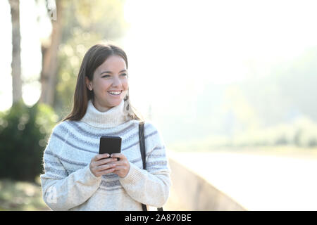 Vorderansicht Portrait eines glücklichen Mädchen mit Pullover Holding smart phone entfernt in einem Park suchen Stockfoto