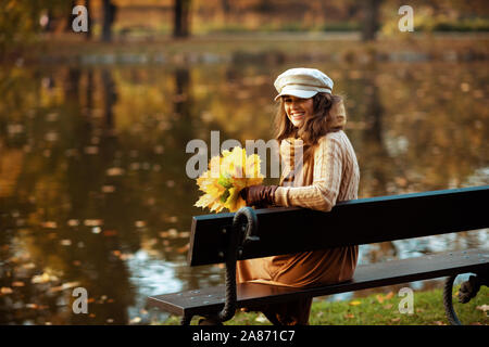 Hallo Herbst. Portrait von Happy trendy Frau, Pullover, Rock, Mütze, Handschuhe und Schal mit gelben Blätter, während auf einer Bank sitzen im Herbst Stockfoto