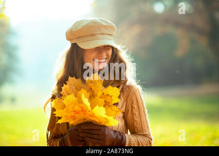 Hallo Herbst. lächelnd modernen mittleren Alter Frau, Pullover, Rock, Mütze, Handschuhe und Schal mit gelben Blätter beiseite Suche im Freien im Herbst Park. Stockfoto