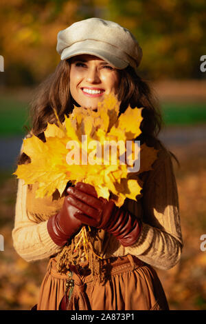 Hallo Herbst. Portrait von Happy trendy 40 Jahre alte Frau in die jacke, rock, hut, handschuhe und Schal mit gelben Blätter in die Ferne schauen, Außenpool Stockfoto