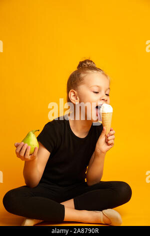 Baby girl Kid essen Vanilleeis in Waffeln Kegel und eine Birne in seine zweite Hand auf gelben Hintergrund mit freiem Text kopieren Raum Stockfoto