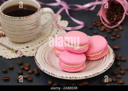 Rosa Makronen sind auf einem Schild auf einem dunklen Hintergrund. Im Hintergrund ist eine Tasse Kaffee Stockfoto