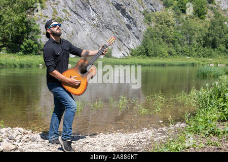 Junge hipster bärtigen Kerl, Mann in einem Gap spielt Gitarre mit abgesenktem Streicher vor dem Hintergrund der hellen Sommer Landschaft Stockfoto