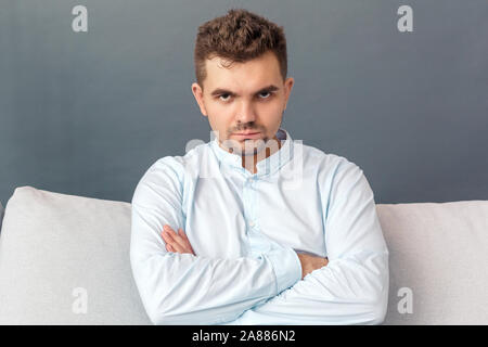 Freestyle. Junger Mann studio Sitzen auf einem Sofa isoliert auf Grau cossed Arme suchen Kamera wütend Stockfoto