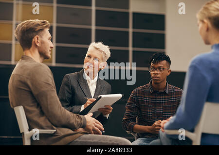 Junge peopple Sitzen auf Stühlen im Kreis und spricht mit den Reifen Psychologe, während Sie Notizen in Dokument während der Therapie Lektion Stockfoto