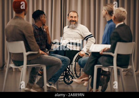 Gerne ältere behinderte Mann sitzt im Rollstuhl und im Gespräch mit den jungen Menschen, die sitzen auf Stühlen im Kreis während der Therapie Lektion Stockfoto