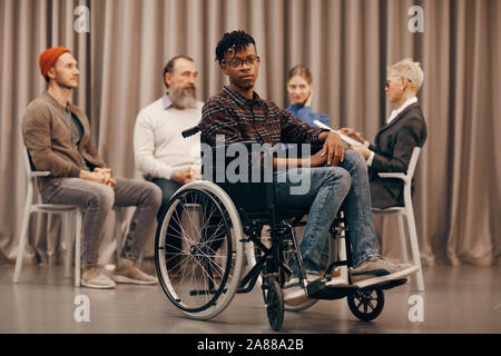 Portrait der Afrikanischen für behinderte Menschen im Rollstuhl an Kamera suchen, während andere Leute etwas diskutieren im Hintergrund Stockfoto