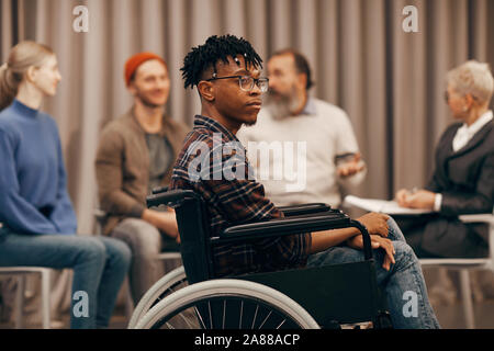 Junge afrikanische behinderte Mann sitzt im Rollstuhl, mit Menschen, die miteinander sprechen, im Hintergrund Stockfoto