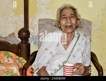 Portrait von lächelnden Active Senior asiatischen und indischen Ethnizität Frau von 82 Jahren sitzen auf Stuhl, eine Tasse Kaffee zu Hause ruht während der Suche Stockfoto