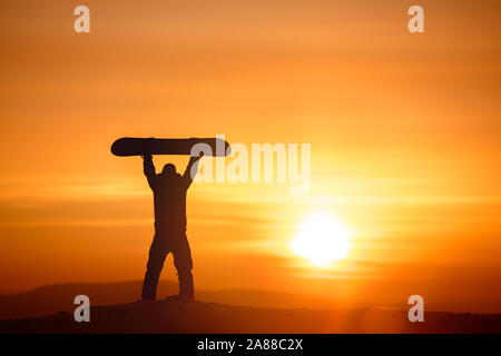 Snowboarder hält Snowboard oberhalb des Kopfes auf Hintergrund der Sonnenuntergang Sonne und Himmel. Ski Konzept Stockfoto