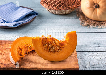 Zwei helle orange Kürbisscheiben mit Samen auf olivenbaum Schneidbrett und blauem Holz- Oberfläche. Kürbis, Tuch und Korb im Hintergrund. Selektive Stockfoto