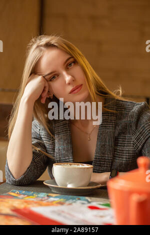 Junge Blondine mit einer Teekanne im Cafe schaut auf Kamera, sehnt sich vermißt, Träume, Hand auf den Kopf Stockfoto