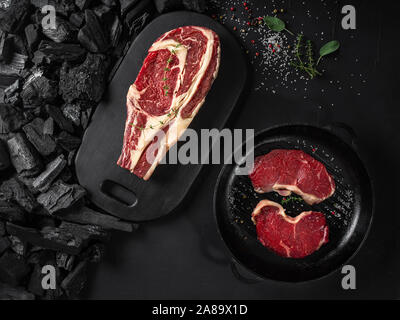 Raw saftiges Steak auf einem dunklen, schwarzen Hintergrund. Frisches Fleisch, das in einem Eisen grillen Pan, Kräuter, Salz und Pfeffer, Kohlen. Zutaten für Cookin Stockfoto