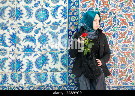 ISTANBUL - Mai 4, 2010: eine junge türkische Frau in hellem Kopftuch steht eine rote Rose mit farbenfroh gemusterten Fliesen Hintergrund. Stockfoto