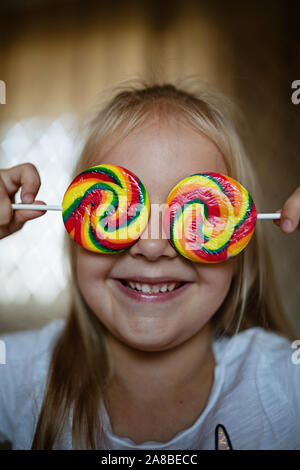 Lustig Kind mit Süßigkeiten Lutscher, Lifestyle Foto von Happy blonde Mädchen essen bunten Zucker lollipop zu Hause Stockfoto