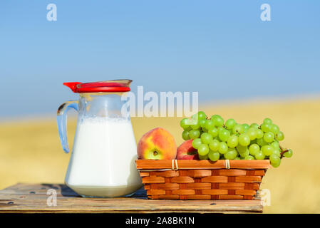 Die ersten Früchte habikkurim in Hebräisch und Milch auf Holztisch. Symbole der jüdische Feiertag - Schawuot. Weintrauben und Pfirsiche auf Korb im Freien auf dem Weizenfeld und blauer Himmel. Stockfoto