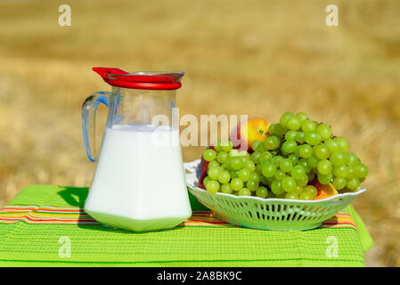 Die ersten Früchte habikkurim in Hebräisch und kanne Milch auf Holztisch mit rustikalem Stoff Handtuch. Symbole Der jüdische Feiertag - Schawuot. Weintrauben und Pfirsiche auf Platte outdoor auf der gold Weizenfeld Hintergrund. Stockfoto