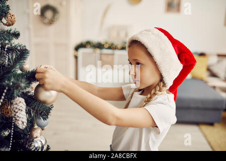 Kleines Mädchen in Santa Hut schmücken Weihnachtsbaum mit Spielzeug für Weihnachten im Wohnzimmer zu Hause Stockfoto