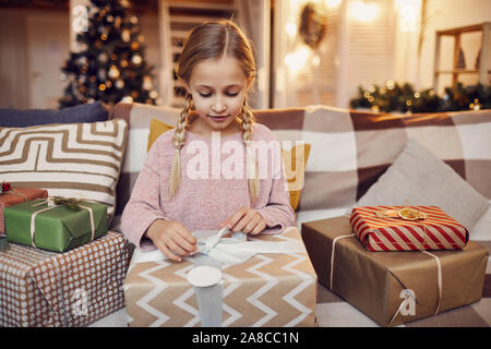 Kleines Mädchen sitzt auf einem Sofa und öffnen großes Weihnachtsgeschenk Sie bekommen vom Weihnachtsmann für Weihnachten zu Hause Stockfoto