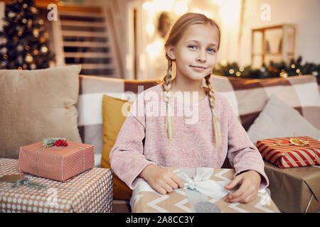 Portrait von niedlichen kleinen Mädchen auf Kamera beim Sitzen auf dem Sofa unter Geschenkboxen und öffnen ihr Weihnachtsgeschenk Stockfoto