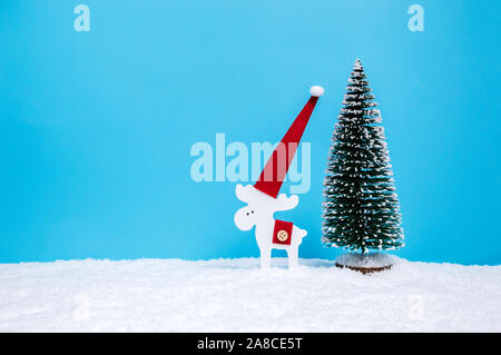 Rentier Deko Figur in Santa's Hut mit Tannenbaum im Schnee vor blauem Hintergrund. Minimalistische Weihnachten Konzept. Frohe Weihnachten greetin Stockfoto