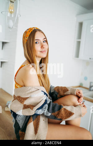 Schönen lächelnden jungen Frau fair lange Haare Mädchen in der gemütlichen Strickjacke auf dem Küchentisch zu Hause sitzen tragen, hellen Innenraum Stockfoto