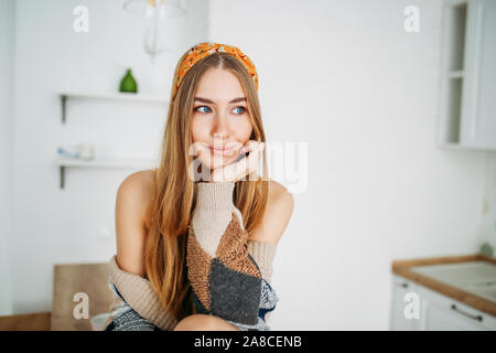 Schönen lächelnden jungen Frau fair lange Haare Mädchen in der gemütlichen Strickjacke auf dem Küchentisch zu Hause sitzen tragen, hellen Innenraum Stockfoto