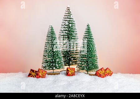 Viele kleine Geschenke unter dem Weihnachtsbaum auf Schnee mit rosa Koralle Hintergrund. Hell erleuchtete Weihnachten Geschenke in Spielzeug Wald. Frohe Weihnachten Grußkarte Stockfoto