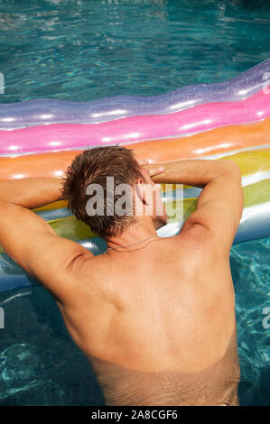 Unerkennbarer Mann ruht seinen Kopf auf einem bunt aufblasbaren Floß, das in einem funkelnden Schwimmbad schwimmt Stockfoto