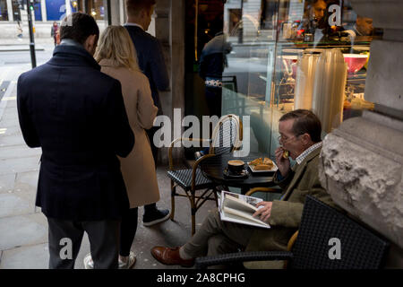 Beim Sitzen auf einem Sitz ausserhalb eines Sandwich Bar auf die Straße, Piccadilly, die eine Warteschlange von Kunden Warteschlange für ihre Mittagessen, als ein anderer Kunde einen Biss in sein Sandwich statt, am 5. November 2019 in London, England. Stockfoto