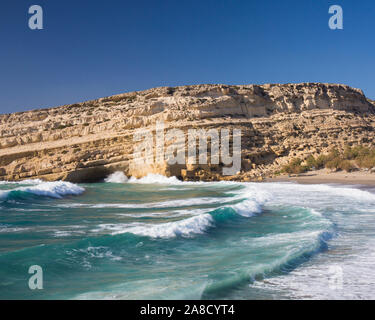 Matala, Heraklion, Kreta, Griechenland. Wellen schlagen der Strand unterhalb Sandsteinfelsen, der ehemaligen Heimat von Cave-dwelling Hippies. Stockfoto