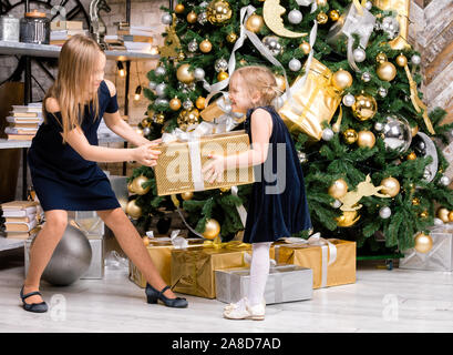 Zwei Schwestern tragen blaue Kleider stehen neben dem geschmückten Weihnachtsbaum ziehen großen goldenen Geschenkkarton verpackt, nicht teilen. ein Geschenk im Boxen da Stockfoto