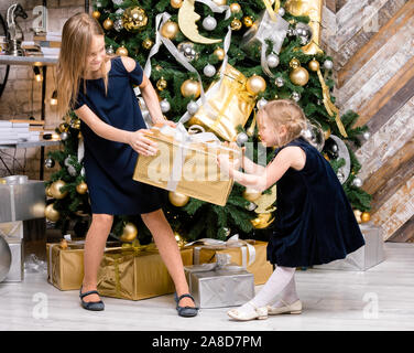 Zwei Schwestern tragen blaue Kleider stehen neben dem geschmückten Weihnachtsbaum ziehen großen goldenen Geschenkkarton verpackt, nicht teilen. ein Geschenk im Boxen da Stockfoto