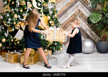 Zwei Schwestern tragen blaue Kleider stehen neben dem geschmückten Weihnachtsbaum ziehen großen goldenen Geschenkkarton verpackt, nicht teilen. ein Geschenk im Boxen da Stockfoto