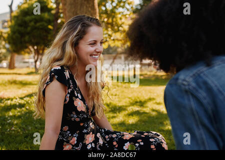 Glücklich lächelnde Blonde schöne junge Frau sitzt mit ihrem weiblichen Freunde Zeit zusammen verbringen in den Park an einem warmen, sonnigen Tag im Sommer Stockfoto