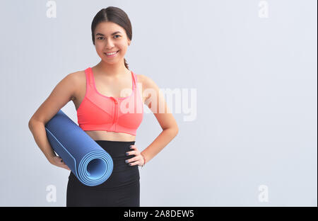 Sportliche lächelnde Mädchen halten Yoga Matte und Übung auf grauem Hintergrund. Sport Konzept Stockfoto