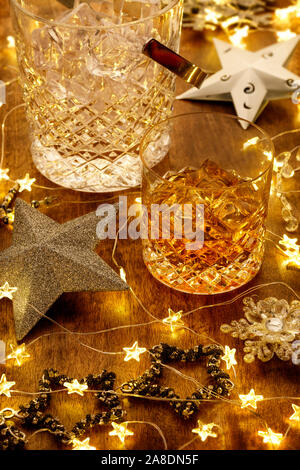 Kristall Glas Whisky und ein Whisky Karaffe mit Weihnachtsbeleuchtung, Kugeln und silbernen Sternen, auf einem Holztisch Stockfoto