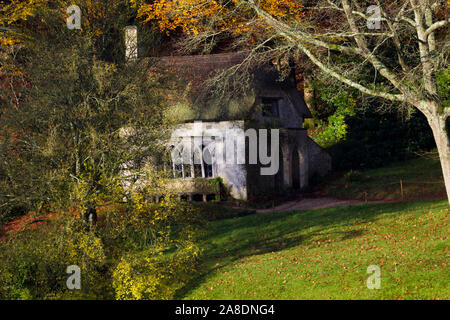 Ein süßes kleines Reetdachhaus in den Wäldern auf der Stourhead Estate. Einen sonnigen, herbstlichen Morgen. Bäume rund um das Haus 2. Stockfoto