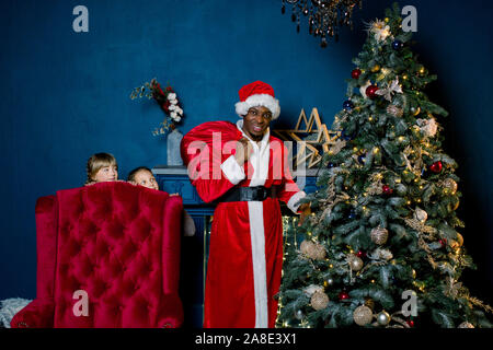 Afrikanische Weihnachtsmann die Geschenke unter dem Weihnachtsbaum in der gemütlich eingerichtete Zimmer, während zwei aufgeregten kleinen Mädchen aus dem roten Stuhl suchen Stockfoto