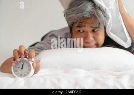 Asiatische Mann mittleren Alters hält eine weiße Wecker und sein Gesicht zeigte, Langeweile und schlechte Gefühl, sein Problem ist eine Schlafstörung. Das Konzept der Suff Stockfoto
