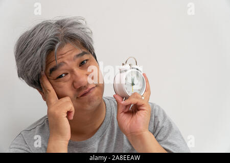 Asiatische Mann mittleren Alters hält eine weiße Wecker und sein Gesicht zeigte, Langeweile und schlechte Gefühl, sein Problem ist eine Schlafstörung. Das Konzept der Suff Stockfoto