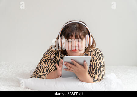 Übergewichtige Frau auf dem Bett und spielen Tablet und das Tragen von Kopfhörern. Das Konzept der komfortable Tag und Rest Stockfoto