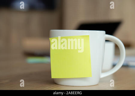 Eine weiße Tasse mit einem leeren gelbe Post-it auf der Schale. Ruht auf dem Schreibtisch und Laptop ist auf der Rückseite platziert. Stockfoto