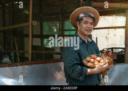 Asiatische Bauer Holding frisches Huhn Eier in Korb und Henne Stand in der Nähe von Henne neben Hühnerfarm. Lächeln, weil mit den Produkten zufrieden aus dem