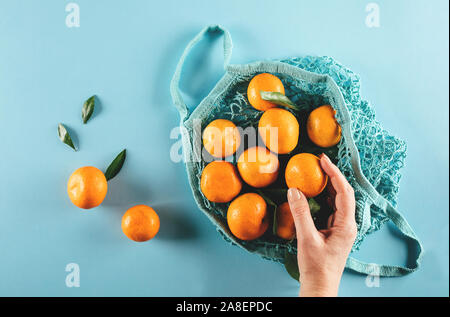 Frau mit einem reifen Mandarine. Mandarinen in mash Shopping Bag auf blauen Tabelle Hintergrund. Null Abfall, umweltfreundliches Konzept. Ansicht von oben, flach Stockfoto